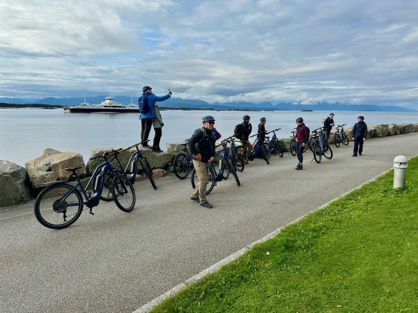 Die E-Bike-Gruppe steht mit den Rädern am Ufer, im Fjord zieht eine Fähre vorbei.