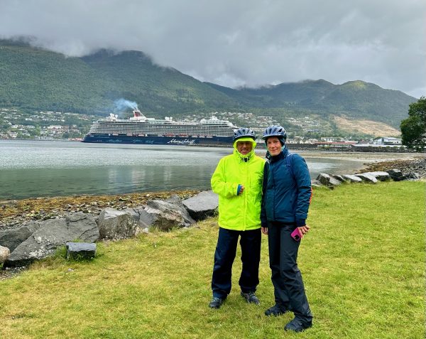 Kathi und Eddy in Regenkleidung mit Fahrradhelmen am Ufer, im Fjord liegt die Mein Schiff 3 vor der wolkenverhangenen Bergkette.