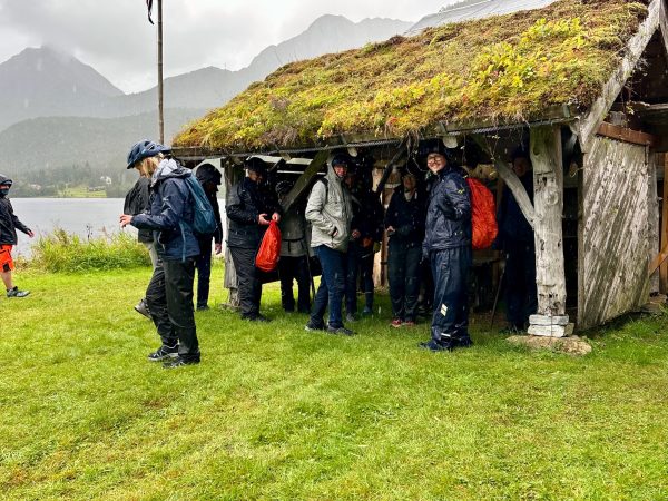 Die Radgruppe steht in Regenkleidung unter dem Vordach einer alten Holzhütte mit Grasdach, im Bild sind Regentropfen zu sehen.