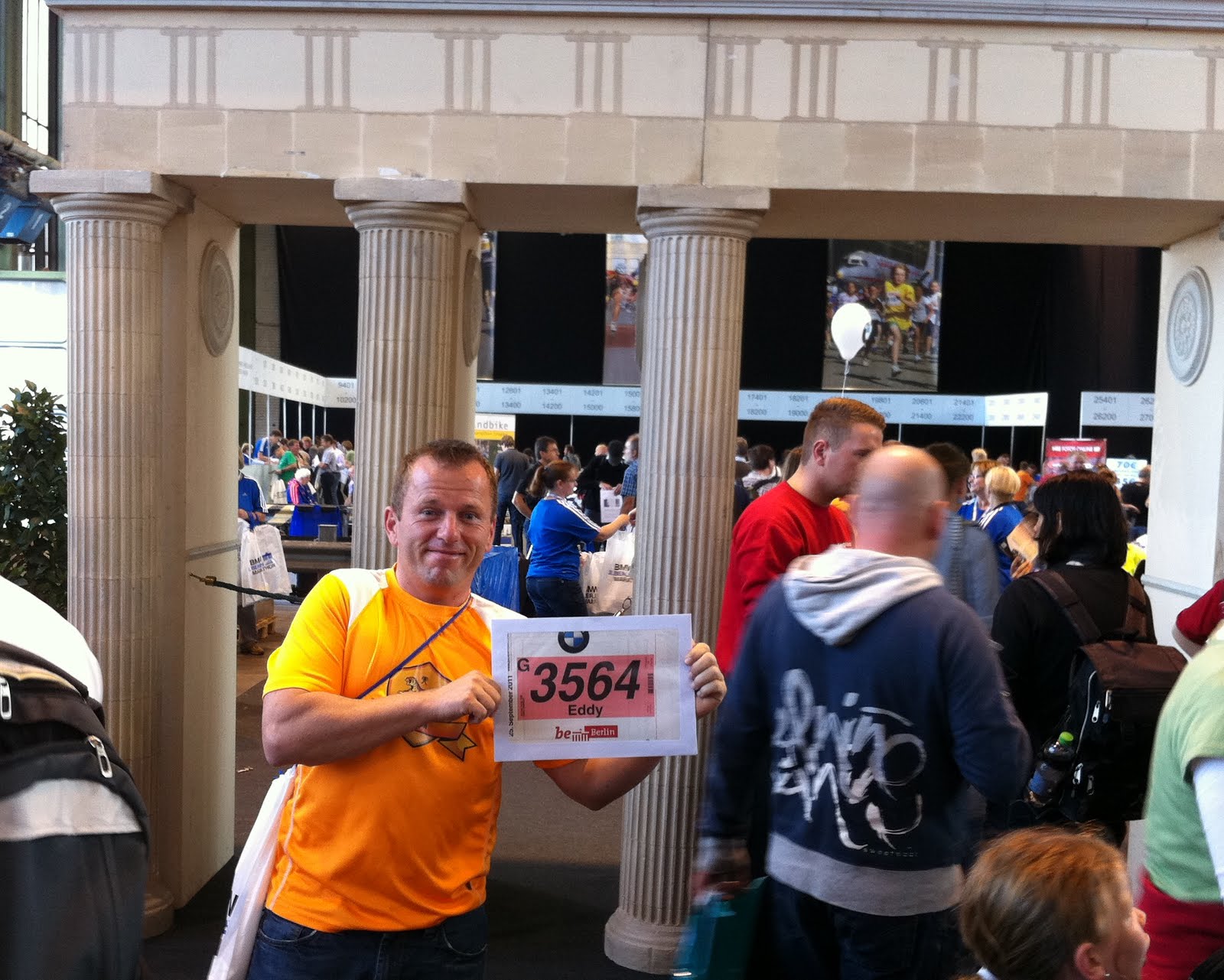Eduard Andrae im orangefarbenen Laufshirt hält auf der Marathon-Messe seine Startnummer G 3564 mit dem Namen Eddy in die Kamera. Hinter ihm stehen Säulen, im Hintergrund drängen sich Besucher in der Messehalle.